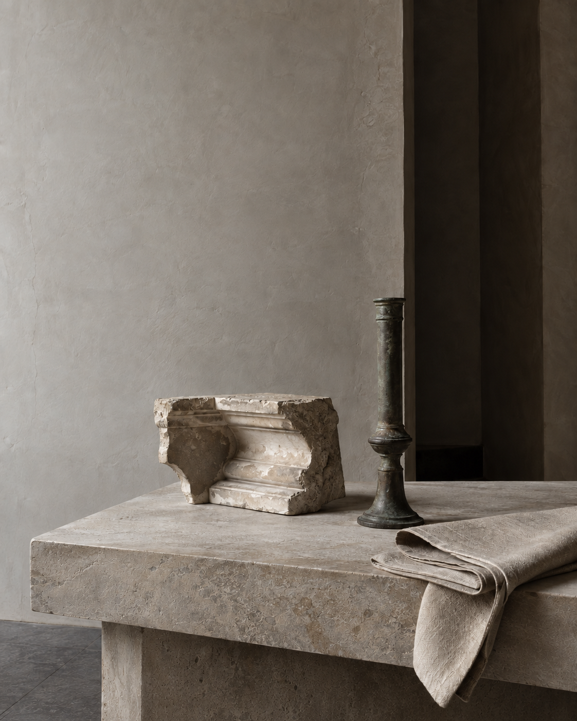 A limestone architectural fragment, aged bronze fitting, and folded linen arranged on a stone worktable, with the linen hanging fully within the frame.